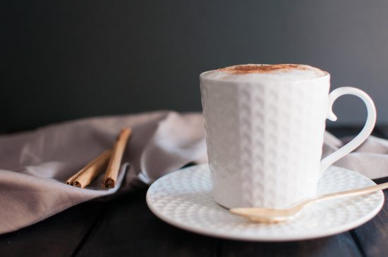A closeup shot of a foamy macchiato in a white ceramic mug. A set of cinnamon sticks sit next to it.