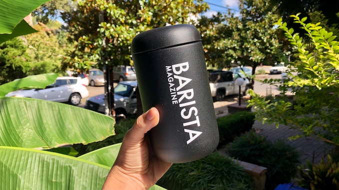 A hand holding a black Fellow takeaway mug with the Barista Magazine logo on it.