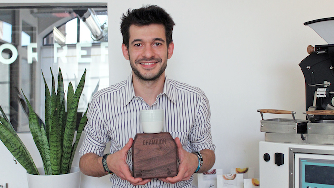 Alessandro is an Italian white male who sits smiling holding a plaque with his latte art champion trophy.