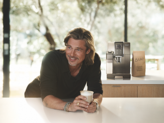 Actor Brad Pitt stands over a counter smiling holding a cup full of a latte. The DeLonghi Espresso Machine is behind him.