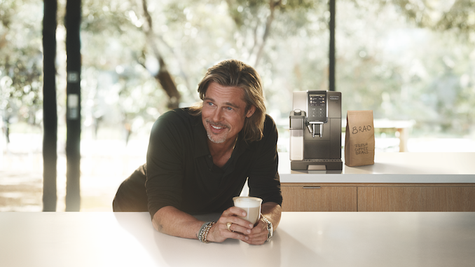 El actor Brad Pitt sonríe sobre un mostrador y sostiene una taza de café con leche en la mano. La máquina de café espresso DeLonghi está detrás de él.