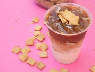 A bright pink background. A plastic cup full of milk layered with coffee and cinnamon toast crunch squares sprinkled across the background.
