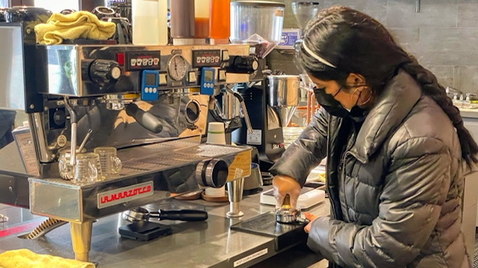 A barista with a grey puffy jacket stands at an espresso machine tamping a shot of coffee.