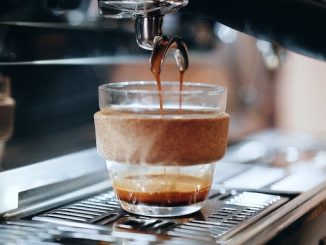 A KeepCup glass ceramic mug stands underneath an espresso machine pouring coffee.