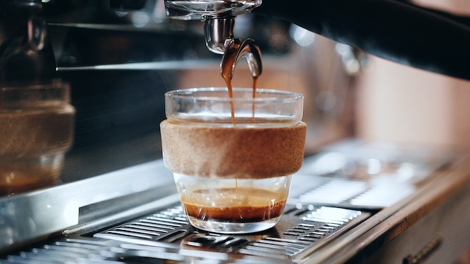 A KeepCup glass ceramic mug stands underneath an espresso machine pouring coffee.