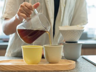 The lower half of a body pouring coffee into mugs on a tray.
