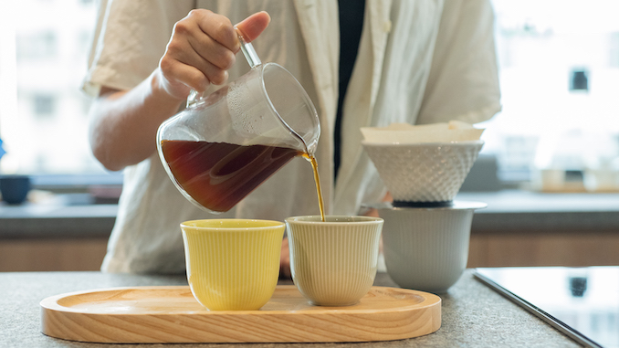 The lower half of a body pouring coffee into mugs on a tray.