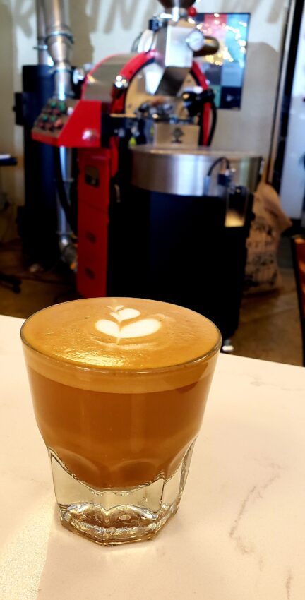 A cortado in a glass on a counter in front of a coffee roaster.