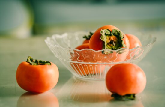 A clear glass bowl is filled with persimmons, some of which have rolled out onto the white tabletop.