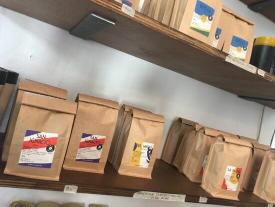 Zwei Holzregale zeigen die bei Brulerie-Moka erhältlichen Kaffeebeutel, die in braunen Kraftpapiertüten verpackt und mit bunten Aufklebern gekennzeichnet sind.
