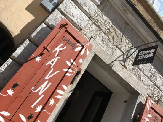 Kapının üzerinde sallanan tabela ve kapının dışında kırmızı panjurlar bulunan Crno Zrno kafeye giriş.