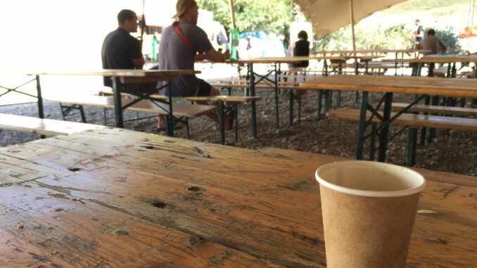 A plain paper coffee cup sits on a wood table under an outdoor festival tent. Two men are seated at another table in the background.