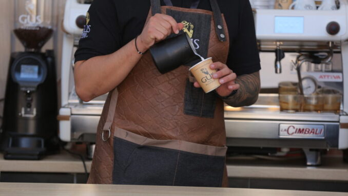 A barista in a leather apron pours steamed milk into a paper cup.