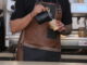 A barista in a leather apron pours steamed milk into a paper cup.