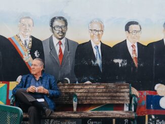 An elderly man sits on a bench, coffee in hand, in front of a mural depicting three men in Miami.