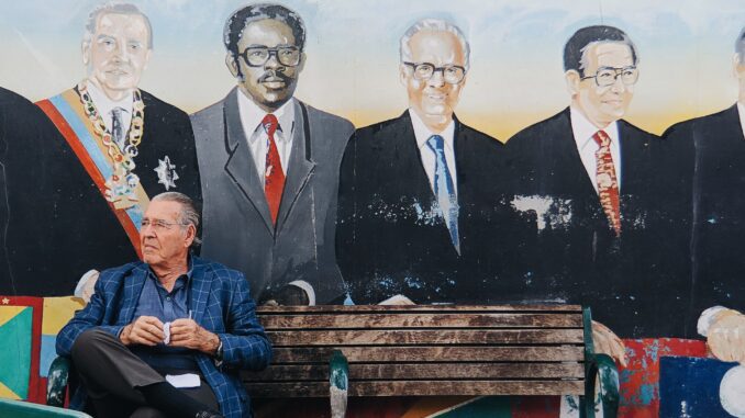 An elderly man sits on a bench, coffee in hand, in front of a mural depicting three men in Miami.