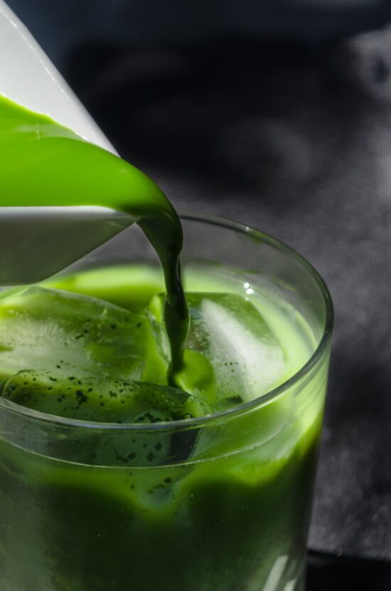 Close up image of matcha green tea pouring from a white porcelain pitcher into a cup of ice. 