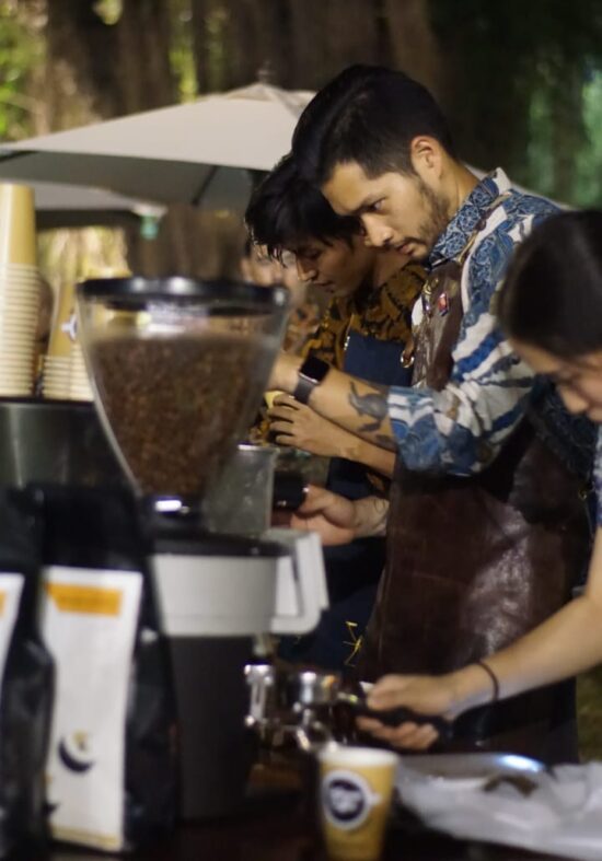 Three baristas stand at an outdoor table with brewing equipment. Jasin is in the middle. A woman in front pulls an espresso shot.