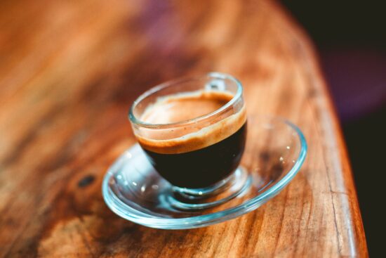 Berrak bir cam espresso fincanı ve tabağındaki bir kafeterya, ahşap bir yüzeyin üzerine oturur.