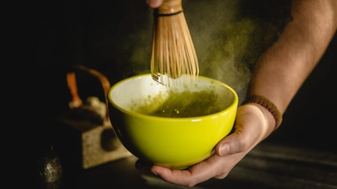 A person holds a yellow bowl of matcha in one hand and a bamboo matcha whisk in the other. They are stirring the matcha with the whisk while tea dust swirls around the top of the bowl.