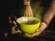 A person holds a yellow bowl of matcha in one hand and a bamboo matcha whisk in the other. They are stirring the matcha with the whisk while tea dust swirls around the top of the bowl.