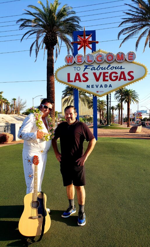 The author stand next to an Elvis impersonator in a white jumpsuit with an acoustic guitar. Behind them is the vintage Welcome To Fabulous Las Vegas! sign with its many neon lights and star at the top.