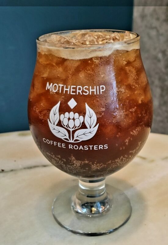 An espresso tonic in a clear glass goblet with the Mothership Coffee Logo printed on front.  The logo is a cascara (or coffee) plant with four leaves wrapped the fruits inside and a tiny diamond above it.