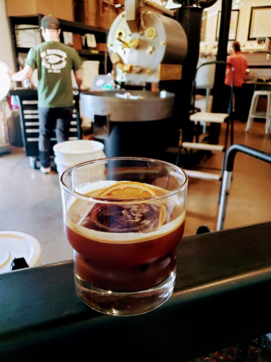 In the foreground, an espresso drink in a small rocks glass, with a slice of orange floating on top.  In the background, a man stands next to a large black coffee roasting machine with a silver tray at the front.