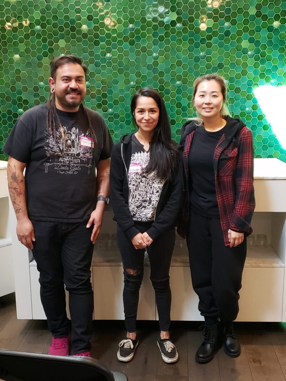 Three people, all with long hair and wearing black, stand together for a photo in front of the green tiled wall at The Crown. They are the Oakland prelims champs.