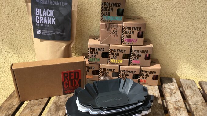 The Comandante upgrades stacked together on a counter: the Red Clix adjuster, spare coffee jars in several colors, large hand crank and small metal coffee trays.