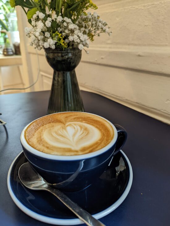 A classic flat white in a navy blue, white rimmed porcelain demitasse cup with handle. The demitasse and a tiny spoon rest on a matching saucer. There is a heart drawn in milk on the flat white. Behind on the plain table top is a vase of white and yellow wildflowers.