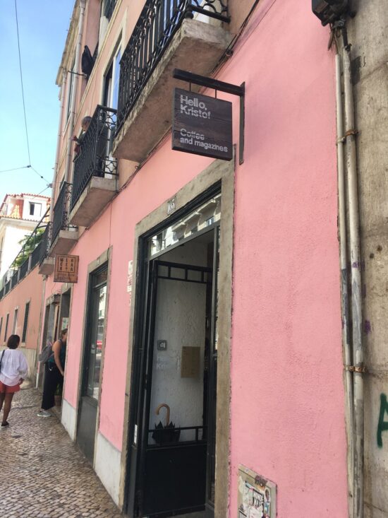 The exterior of Hello, Kristof, at the front of a pink building. The sign is small and black, and swings above the door.