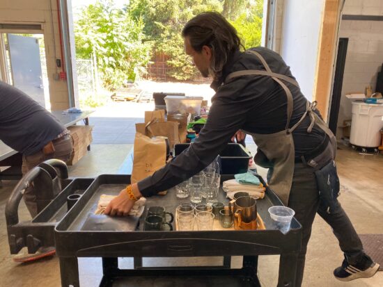 A long-haired barista in a handmade apron prepares his coffee cart for presentation. One the plastic cart are scales, coffee cups, spoons and other barista tools for a successful coffee presentation.