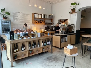 Wnętrze Isla Coffee w Berlinie.  Bar z wbudowanymi półkami na wino.  Półki na tylnej ścianie mieszczą olbrzymie torby z kawą.  Na blacie stoi wybór ciastek, a w rogu stoi ekspres do kawy.
