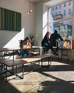 Two cafe-goers sit at a bar-height table by a large window.  Another customer sits alone at a picnic-style table inside.