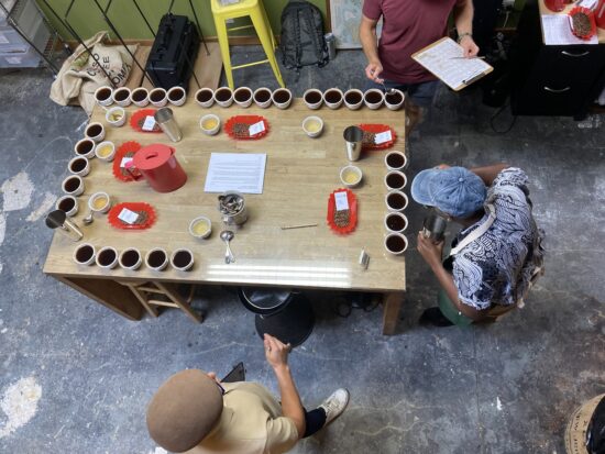 Jay and two other people stand around a cupping table with many cups and bean trays.