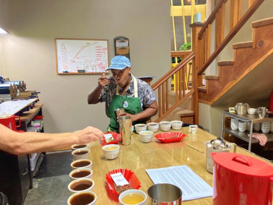 Jayy Terrell sips from spoon during a cupping. Jay wears a green apron and patterned shirt. On the table are metal tumblers and ceramic cups. Someone's arm is reaching a spoon over a cup.