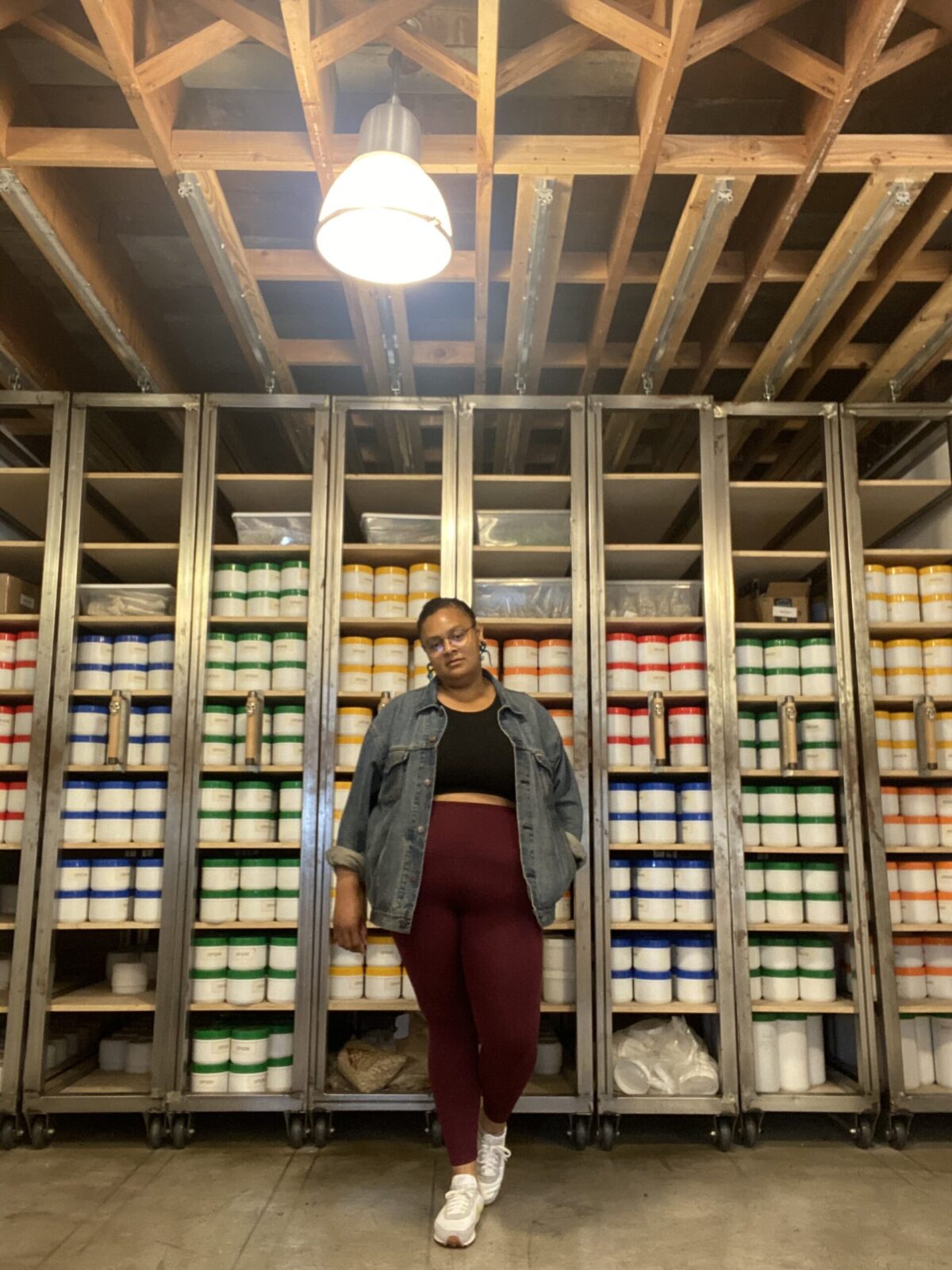 Porttia poses for the camera, one leg bent, In front of shelves of dry goods. In the foreground is a ceiling lamp.