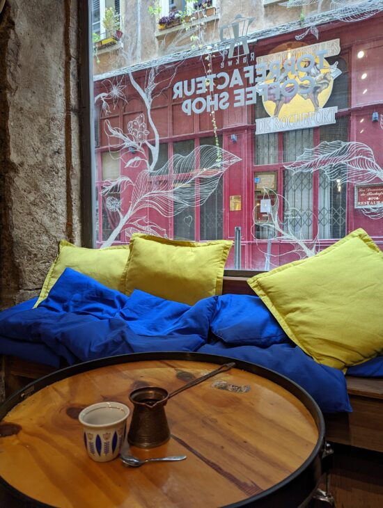 A cozy corner of Rakwé cafe. There is a wooden drum-shaped coffee table in the middle of a set of couches covered in thick cushions. On the table is a porcelain tulip-shaped espresso cup with painted flowers and leaves. Next to it is a traditional Lebanese coffee maker; it is bronze colored, shaped like an upside-down bell, with a long metal handle on top, similar to the handle of a large spoon. 