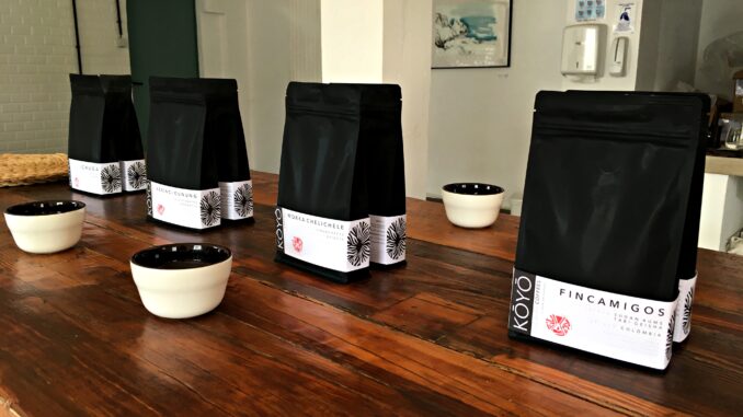 A wooden table with porcelain cups laid out in front of coffee bags. The bags each represent a different coffee that will be tasted.