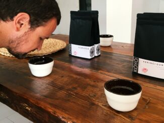 A bearded man leans over a porcelain coffee sup to smell coffee as it brews in the cup for a cupping.
