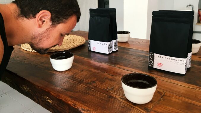 A bearded man leans over a porcelain coffee sup to smell coffee as it brews in the cup for a cupping.