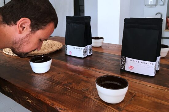 Derselbe Holztisch, Porzellantassen und Kaffeebeutel.  Ein Mann beugt sich über Kaffee, um zu riechen, während er in der Tasse aufbrüht und sich darauf vorbereitet, probiert zu werden. 