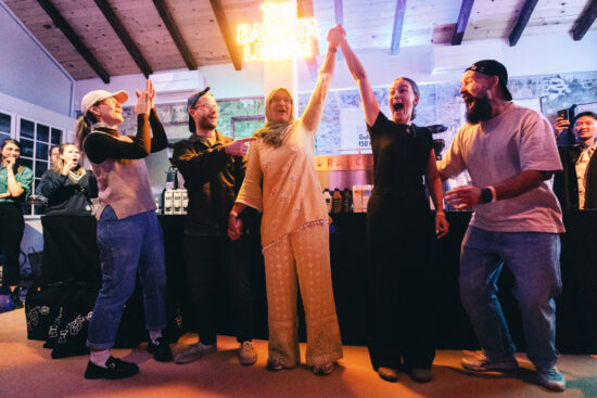 Five people on the Bolivian team. One woman in all black, one on in peach clothes, wearing a headscarf. They are raising their arms together. Another woman in a baseball cap and ponytail is clapping. Two men are laughing and pointing at each other.