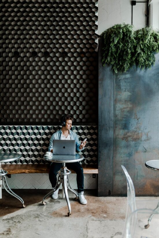 A person by themselves at a table a cafe with a phone and laptop computer.