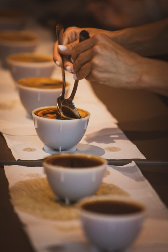 A person uses two spoons to break the crust of coffee grounds on top of a white porcelain cup of coffee during cupping.