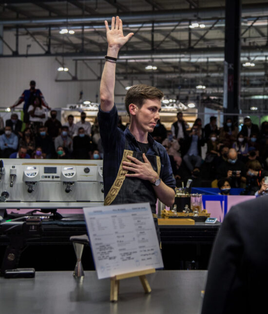 A competition barista raises up his arm in the air, other hand on his chest, as time is called on his presentation.