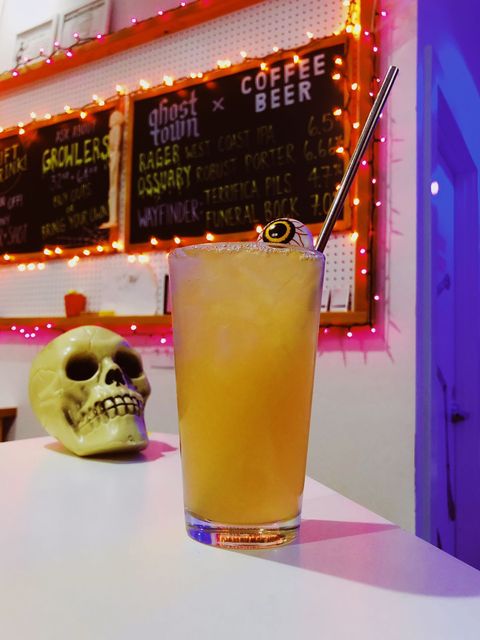 In the background sits a skull on a white countertop. In the foreground is an iced apple cider drink with a metal straw and a floating eye ball on top.