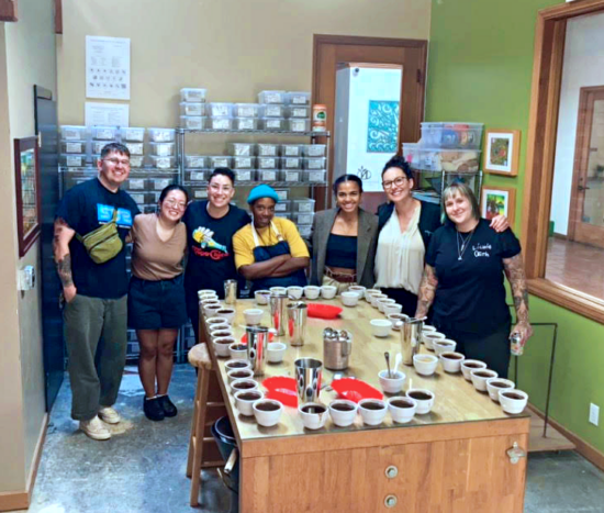 Jay sits at a cupping table, smiling with arms folded. A group of six people surround them, smiling for the photo. On the cupping table are about 30 cups, metal tumblers, spoons, coffee trays and a pitcher. 