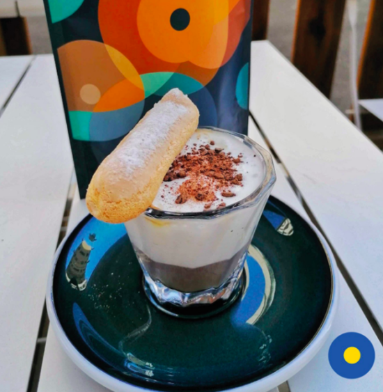 On a saucer, a small decorated glass filled with the espresso tiramisu drink. The espresso sits on the bottom while steamed milk floats on top, dusted with chocolate powder. On the ledge of the cup, a ladyfinger cookie is rested.